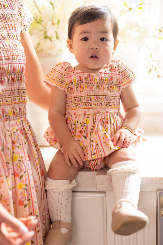 An infant wearing the Ivy City Co Baby Marlow Romper in Peach Handpainted Flora Stripe with white knee-high socks sits on a counter next to an adult dressed in a coordinating outfit.