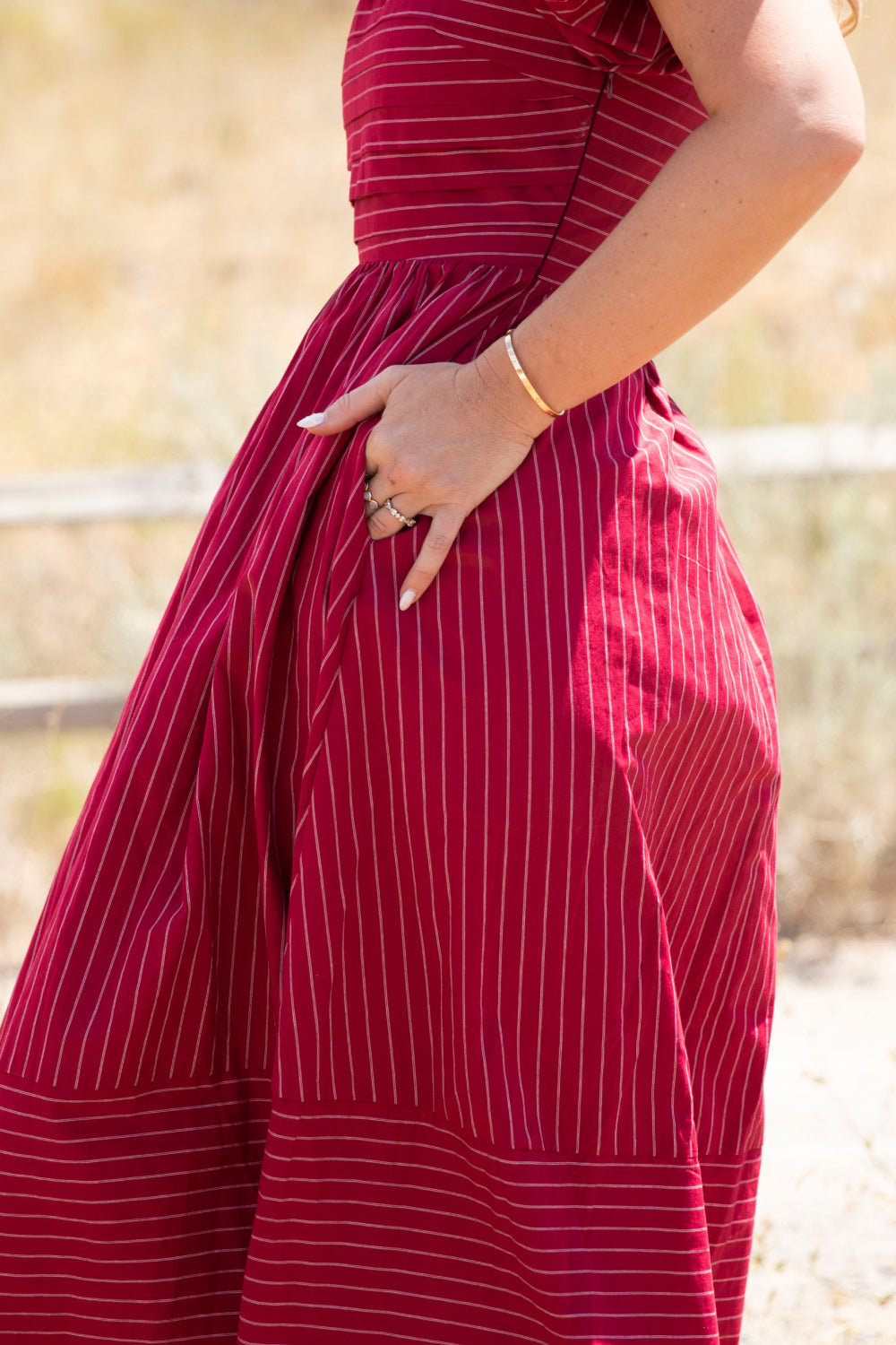 A person wearing the Ivy City Co Julie Dress in Cranberry Pinstripe, which features puff sleeves and a smocked bodice, stands outdoors with one hand on their hip. Only their torso and arm are visible against a blurred natural backdrop.