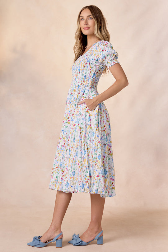 A woman stands against a soft beige background wearing the Amelie Dress in Floral Fields by Ivy City Co—a light blue, floral-print, smocked midi dress with short puff sleeves and pockets—paired with blue heeled shoes adorned with bows.
