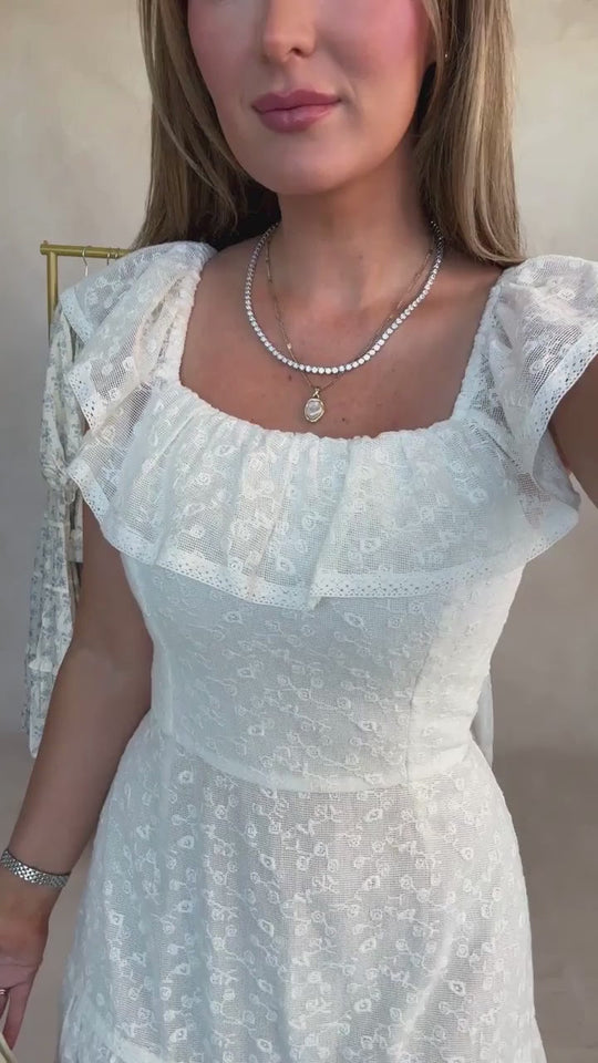 A woman wears the Alaina Dress in Ivory by Ivy City Co—featuring a delicate lace-patterned, smocked bodice, ruffled sleeves, and a square neckline—styled with layered necklaces as she stands before a neutral background.