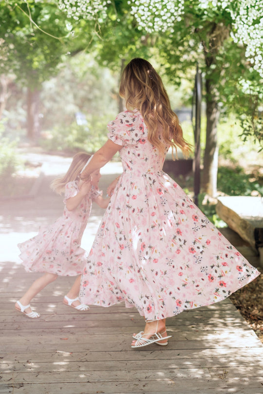 A woman and a young girl twirl together on a sunlit wooden path surrounded by greenery, both wearing the Ivy City Co Sophia Dress in Roselyn Pink.