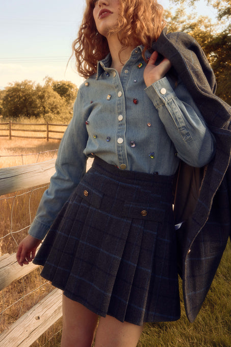 A woman with curly hair wears the Ivy City Co Kinley Skirt in Charcoal Plaid, paired with a denim shirt and holds a matching wool blend jacket over her shoulder, standing by a wooden fence in a sunlit rural field.