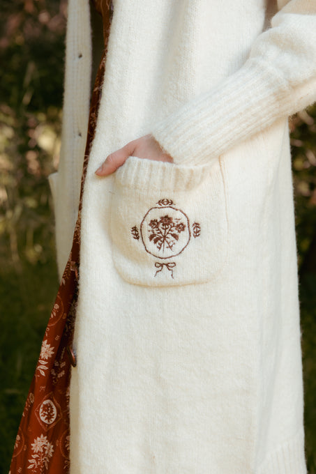 A person wears the Teresa Cardigan in Cream by Ivy City Co, featuring brown floral embroidery and a front pocket, paired with a brown patterned dress. The stylish outfit is maternity nursing friendly in a green outdoor setting.