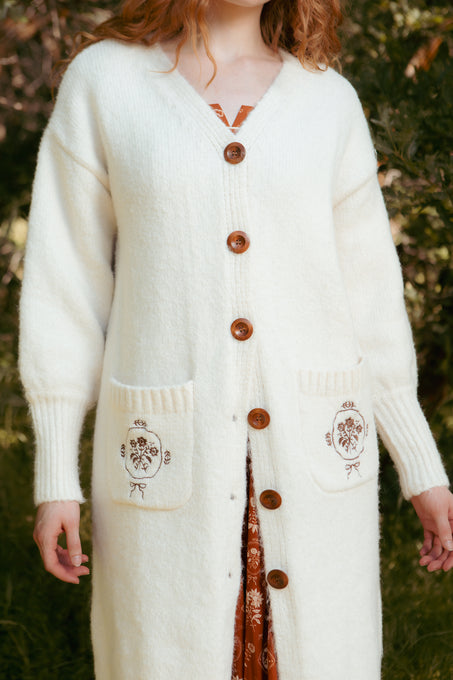 A woman stands outdoors wearing the Ivy City Co Teresa Cardigan in Cream, featuring button-up style and floral-embroidered pockets, layered over a brown patterned dress. The nursing-friendly outfit is shown cropped at her chin and below the knees.