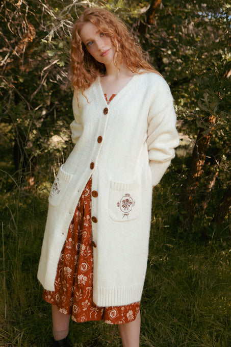 A woman with curly red hair stands outdoors among trees, wearing the Ivy City Co Teresa Cardigan in Cream over a rust floral dress. She looks at the camera, smiling gently in this maternity and nursing-friendly outfit.