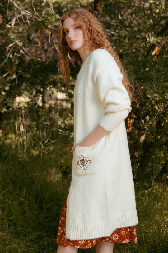 A woman with long curly red hair stands outdoors on grass, wearing the Teresa Cardigan in Cream by Ivy City Co over a rust-patterned dress. This maternity and nursing-friendly cardigan features an embroidered pocket, with trees and foliage behind her.