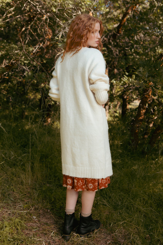 A woman with wavy red hair stands outdoors, back turned, wearing Ivy City Co’s Teresa Cardigan in Cream over a brown patterned dress with black boots, surrounded by green trees and grass.