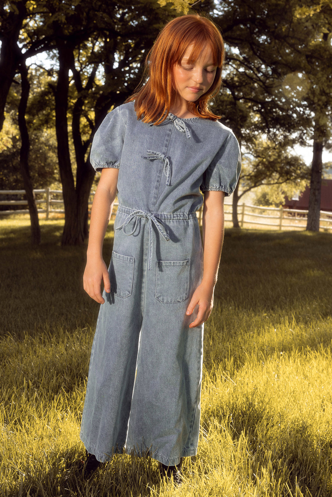 A young girl with red hair stands on grass, wearing the Ivy City Co Mini Dallas Jumpsuit in Denim, featuring puff sleeves and front pockets. Trees and a wooden fence are visible in the sunny background—perfect for a mommy and me day out.