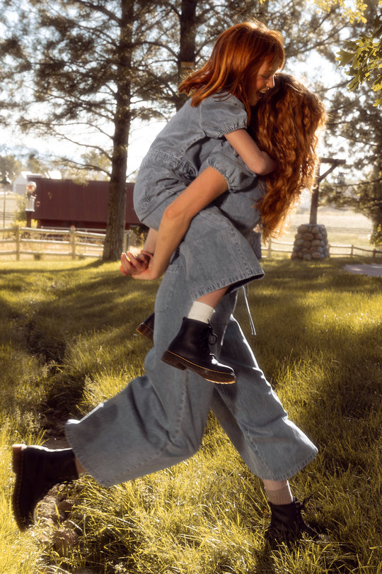 Two people with long red hair, dressed in Ivy City Cos Mini Dallas Jumpsuit in Denim and black boots, share a playful piggyback ride through sunlit grass outdoors—a perfect mommy and me moment by trees and a wooden fence.