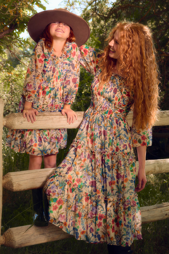 A woman and young girl with long red hair wear matching Ivy City Co Mini Selena Dresses in Vivid Meadow, featuring tiered ruffle skirts. The girl smiles under a wide-brimmed hat as warm sunlight washes over them by a wooden fence outdoors.