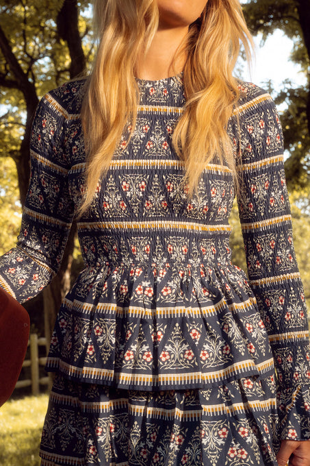 A woman with long blonde hair stands outdoors in sunlight, surrounded by trees and green foliage, wearing the Ivy City Co Greer Dress in Navy Fleur with a smocked bodice.