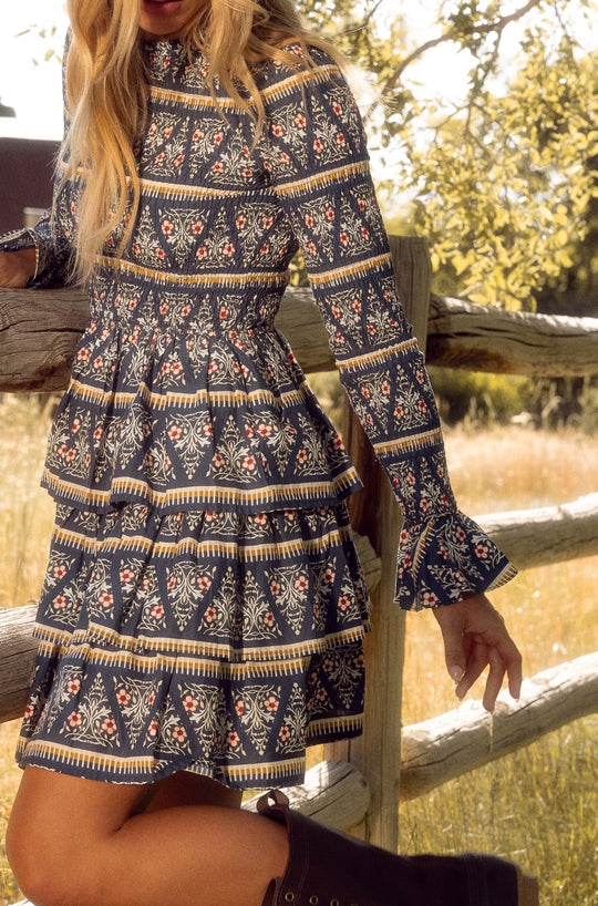A woman with loose blonde hair and brown boots stands by a wooden fence in the sun, wearing the Ivy City Co Greer Dress in Navy Fleur—a smocked-bodice mini dress with long sleeves.