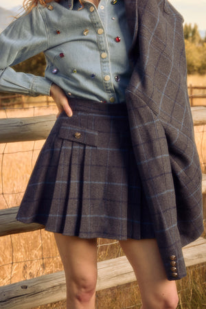 A person stands by a wooden fence in a field, wearing the Ivy City Co Kinley Skirt in Charcoal Plaid, with a matching jacket over one shoulder and a denim shirt with colorful buttons. Only the lower half of the face and body are visible.