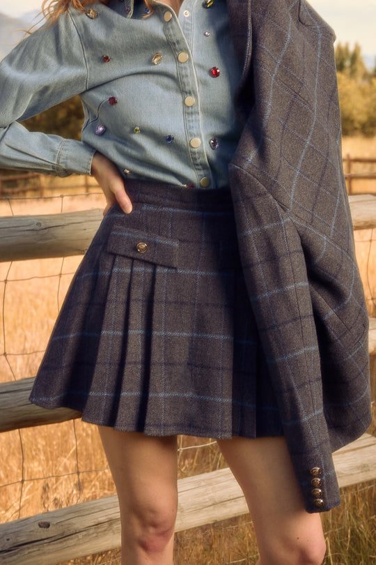 A person stands by a wooden fence in a field, wearing the Ivy City Co Kinley Skirt in Charcoal Plaid, with a matching jacket over one shoulder and a denim shirt with colorful buttons. Only the lower half of the face and body are visible.