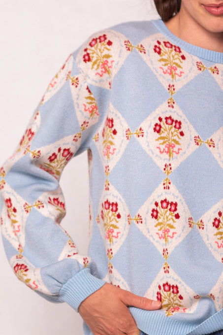 A person wearing the Vivienne Sweater in Mosaic Bloom by Ivy City Co—a light blue sweater with a diamond pattern and red and yellow floral designs; only their upper body, part of an arm, and hand are visible.