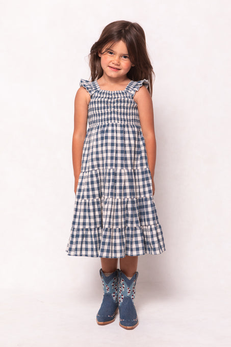 A young girl with long brown hair smiles while wearing the Ivy City Co Mini Hailey Dress in Indigo Gingham, featuring a smocked bodice, and blue floral cowboy boots, standing against a plain light background.