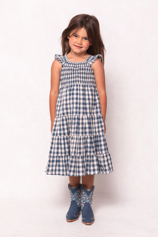 A young girl with long brown hair smiles while wearing the Ivy City Co Mini Hailey Dress in Indigo Gingham, featuring a smocked bodice, and blue floral cowboy boots, standing against a plain light background.