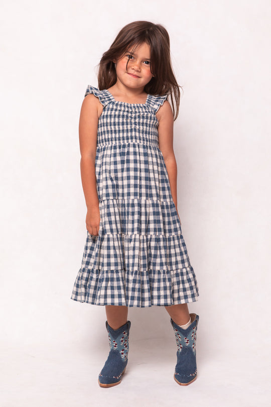 A young girl with long brown hair wears the Ivy City Co Mini Hailey Dress in Indigo Gingham and blue floral boots, standing and smiling with her head slightly tilted against a light background.