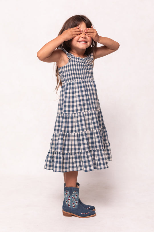 A young girl smiles with her hands over her eyes, wearing the Ivy City Co Mini Hailey Dress in Indigo Gingham, featuring a smocked bodice and midi length, plus blue embroidered boots, against a plain background.