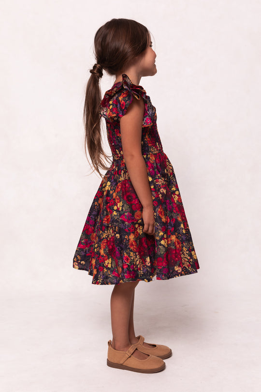 A young girl with long brown hair in a ponytail stands in profile, wearing Ivy City Co’s Mini Hattie Dress in Night Garden with ruffled sleeves and tan Mary Jane shoes, set against a light, plain background.
