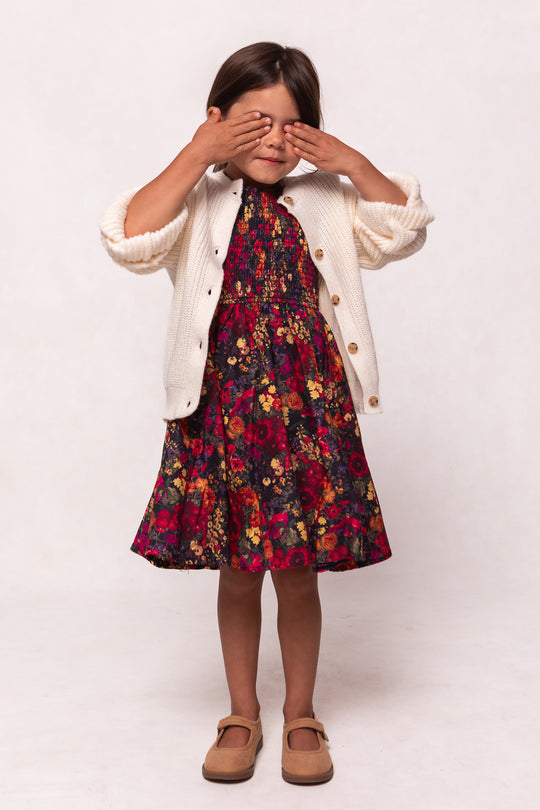 A young girl wears the Ivy City Co Mini Hattie Dress in Night Garden, paired with a cream cardigan and tan shoes, standing against a plain light background with her eyes covered by her hands.