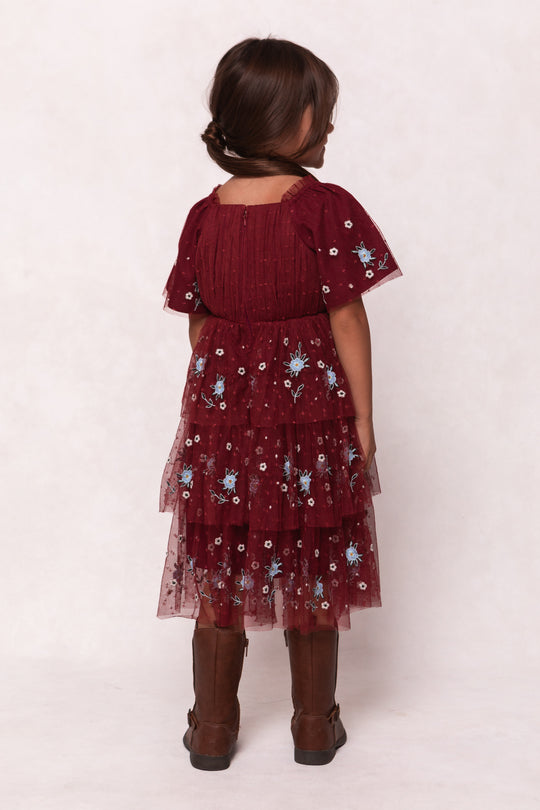 A young girl faces away, wearing the Ivy City Co Mini Mabel Dress in Burgundy with floral embroidery and a tiered tulle skirt. Her loose braid and brown knee-high boots stand out against a simple, light background.