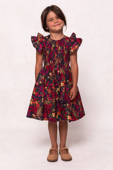 A young girl with brown hair smiles against a plain background, wearing the Ivy City Co Mini Hattie Dress in Night Garden—a dark floral midi dress with ruffled sleeves—perfect for a stylish girls look or mommy and me matching outfit.