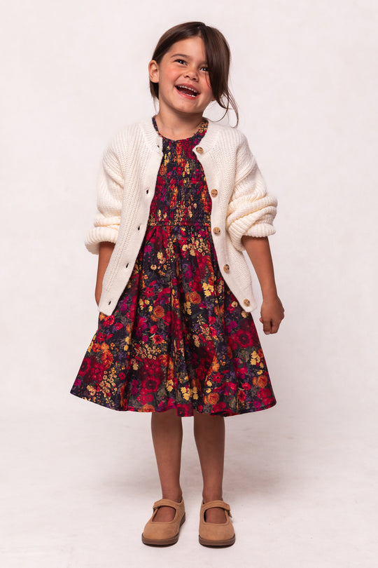 A young girl with brown hair smiles while wearing the Ivy City Co Mini Hattie Dress in Night Garden, featuring a red and yellow floral pattern, paired with a white knit cardigan and tan shoes.