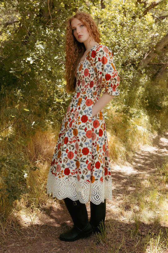 A young woman with long curly red hair stands on a sunlit, leafy path in the Ivy City Co Teagan Dress in Sepia Flower—a floral, maternity-friendly midi—paired with black tights and shoes. She looks calmly at the camera, surrounded by greenery.