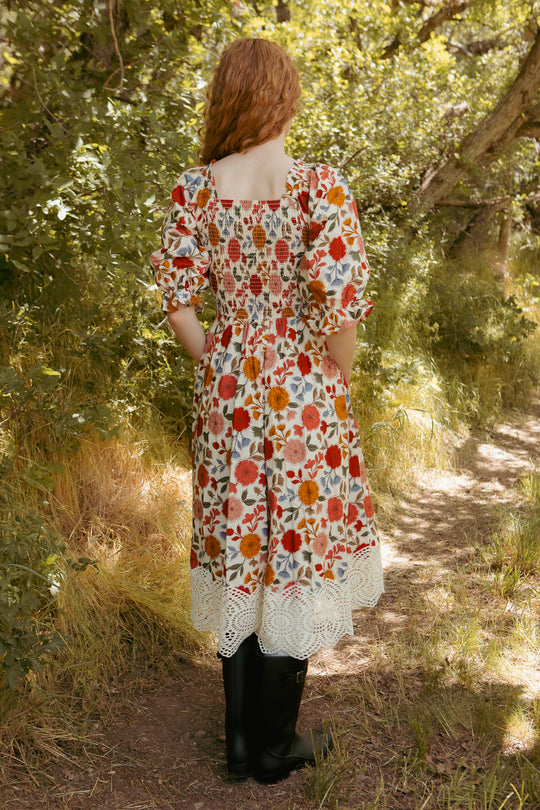 A woman with long, curly red hair stands on a sunlit forest path, wearing the Teagan Dress in Sepia Flower by Ivy City Co—a lace-trimmed, floral midi dress that’s maternity friendly and perfect for any occasion.