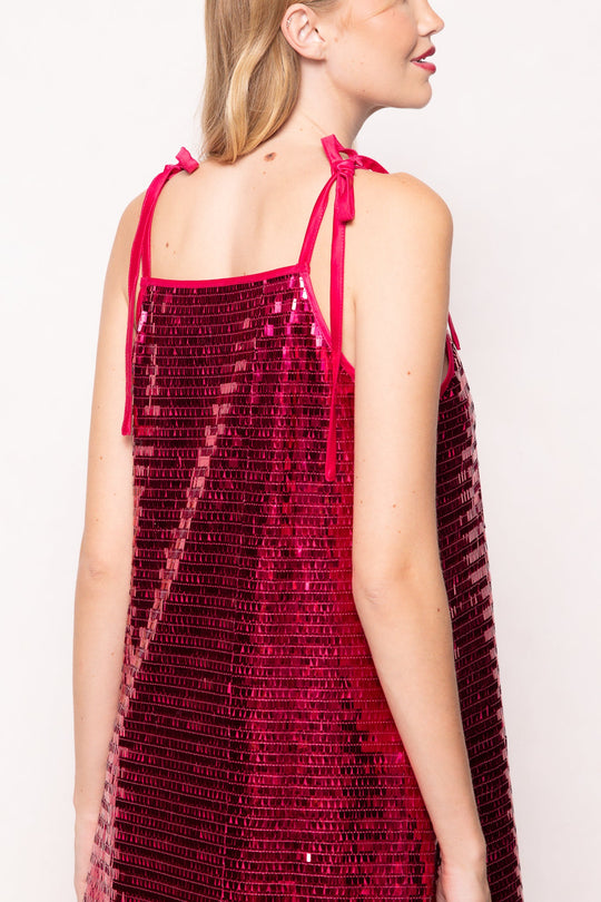 A person wearing the Zelda Dress in Fuchsia Shingle Sequin by Ivy City Co—a sleeveless party dress with ribbon ties on the shoulders—shown from the back and side against a plain light background.
