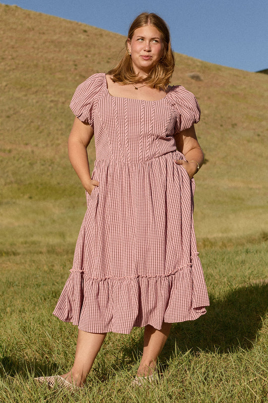 A woman stands on grass in front of a hill, wearing the Ivy City Co Monica Dress in Pink & Brown Gingham, which features short sleeves and a ruffled hem, looking slightly to the side.