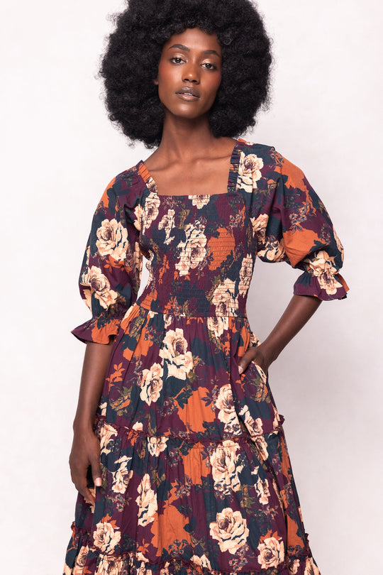 An effortlessly stylish woman with an afro hairstyle wears the Ivy City Co Madeline Dress in Equinox Rose—a long, floral midi dress with puffed sleeves and a square neckline, showcasing vibrant orange, burgundy, and cream hues. Maternity friendly, she stands confidently with one hand on her hip against a light background.