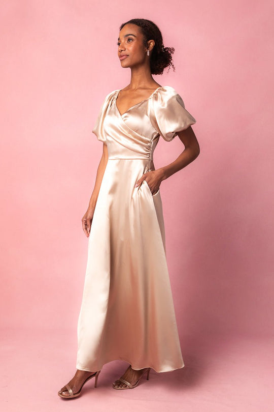 A woman poses against a pink background wearing the Tessie Dress in Champagne by Ivy City Co, featuring puff sleeves and a wrap-style satin bodice.