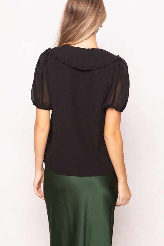 A woman with long, straight hair is shown from the back against a plain light background, wearing the Andrea Blouse in Black Chiffon by Ivy City Co paired with a green skirt.