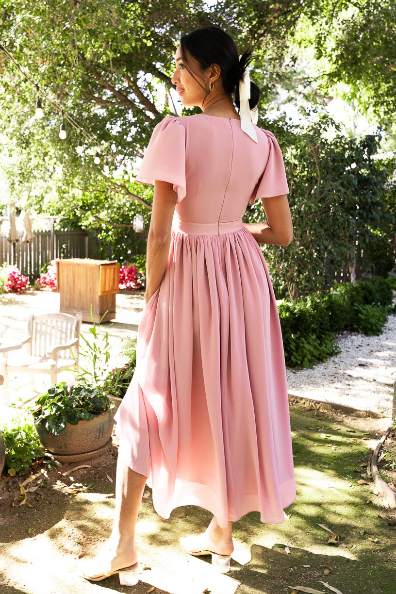 A woman wearing the Abbie Dress in Blush by Ivy City Co, featuring flutter sleeves, stands outdoors in a sun-dappled garden with her back turned. A white ribbon adorns her hair and she pairs the dress with light-colored flats. Elegant trees and garden furniture complete the scene, highlighting this modest bridesmaid dress.
