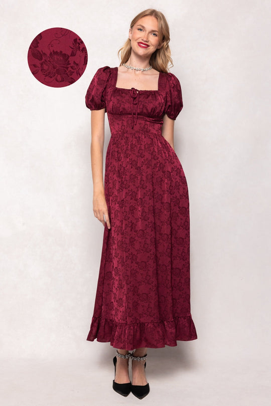A woman smiles while wearing the Amira Dress in Red by Ivy City Co—a burgundy floral-patterned jacquard maxi dress featuring puff sleeves, a square neckline, and a ruffled hem. She pairs the dress with black heels. A circular inset highlights the dress’s detailed fabric.