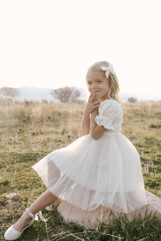 A young girl wears the Ivy City Co Mini Ballerina Dress in White, featuring a delicate tulle skirt and bow, as she sits on the grass in a sunlit field, smiling shyly with her hands near her face. The warm sunlight creates a gentle glow in the background.