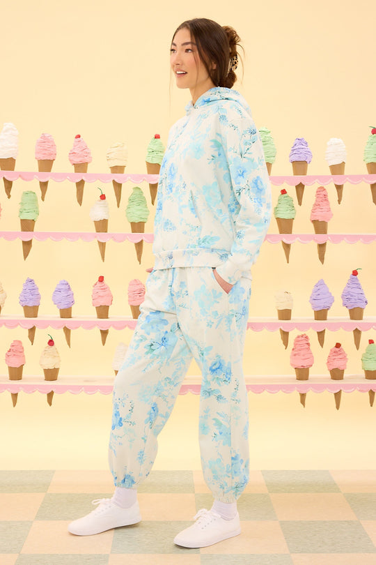 A woman wearing the Ivy City Co Kian Jogger Sweatpants in Blue Aquarelle Fleur and white sneakers stands in front of a pastel ice cream cone display on a pale checkered floor, with a light yellow background.