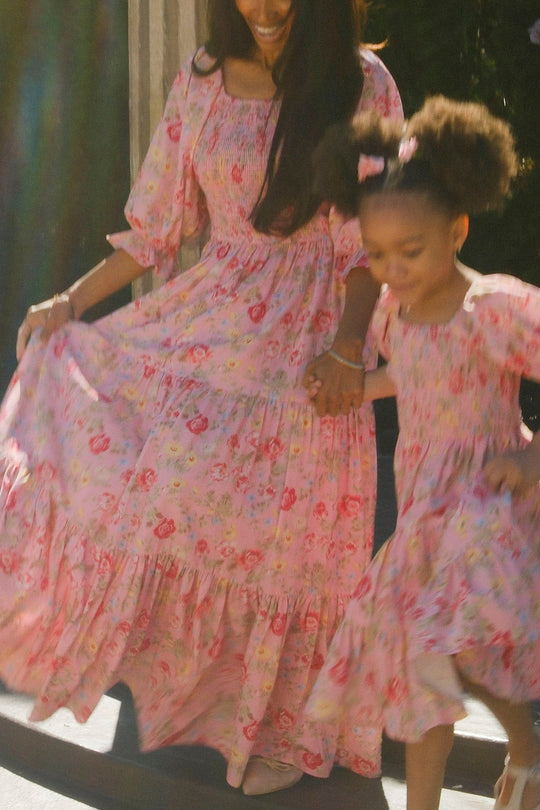 A woman and a young girl twirl hand in hand outdoors, both beaming in matching Ivy City Co Madeline Maxi Dresses in Pink Vintage Poppies. Sunlight bathes the scene in warmth, highlighting the dresses’ charming floral pattern and nursing-friendly design that combines style with practicality for mothers.