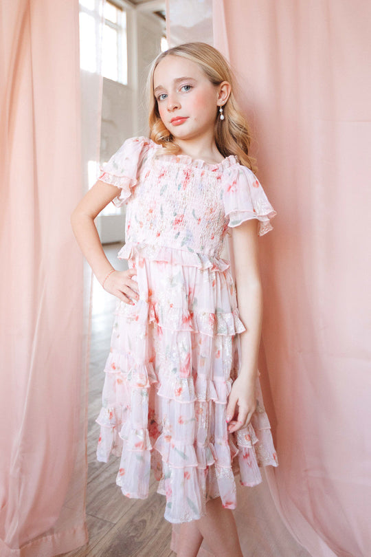 A young girl with soft blonde curls wears the charming Mini Winnie Dress in Pink Floral by Ivy City Co, a ruffled dress featuring pink and white hues. She stands indoors against pale pink curtains, one hand on her hip, gazing confidently at the camera.