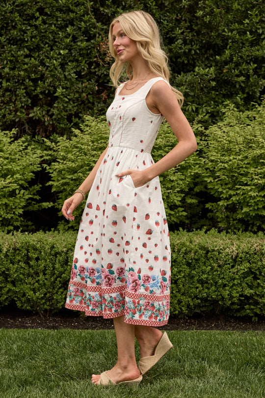 A woman wearing the Sadie Dress in Strawberry Vintage Tier by Ivy City Co—a sleeveless cotton summer dress featuring a strawberry and floral print—stands on grass, posing in front of green bushes. She pairs the look with beige wedge shoes and has long blonde hair styled in loose waves.