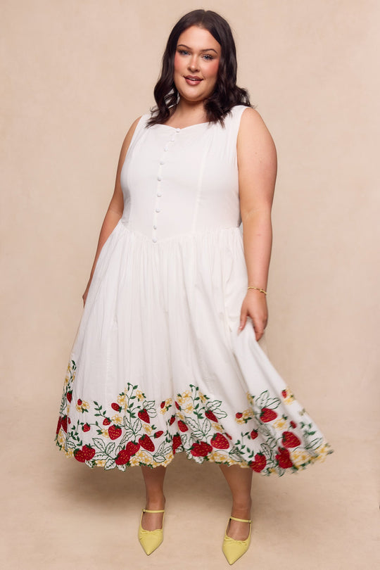 A woman stands smiling against a beige background, wearing the Clarissa Dress in Strawberry by Ivy City Co—a sleeveless white cotton graduation dress featuring colorful floral embroidery at the hem. She pairs it with yellow pointed-toe shoes and has wavy, shoulder-length dark hair.