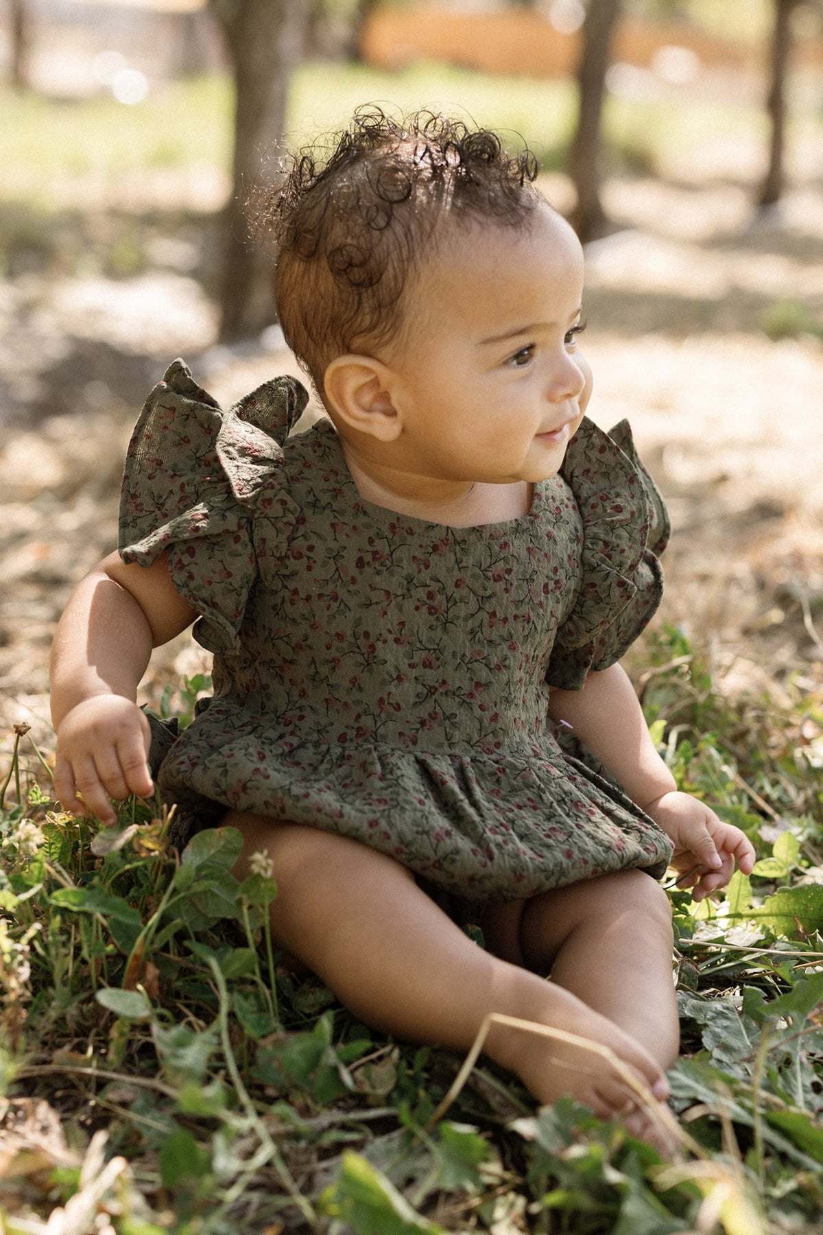 An infant in the Ivy City Co Baby Hattie Romper in Woodland Floral, featuring flutter sleeves, sits on the grass outdoors amidst sunlight and trees, gazing to the side with a gentle smile.