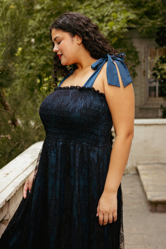 A woman with curly dark hair stands outdoors near greenery and a white railing, gently smiling as she looks down, wearing the Ivy City Co Isadora Dress in Navy & Black—a maternity-friendly style with ribbon straps.