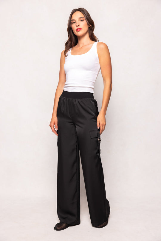 A woman with long brown hair and a neutral expression stands against a plain background, wearing the Ivy City Co Shay Satin Pants in Black—loose fit cargo pants featuring an elastic waistband and side pockets—paired with a white sleeveless tank top and black shoes.