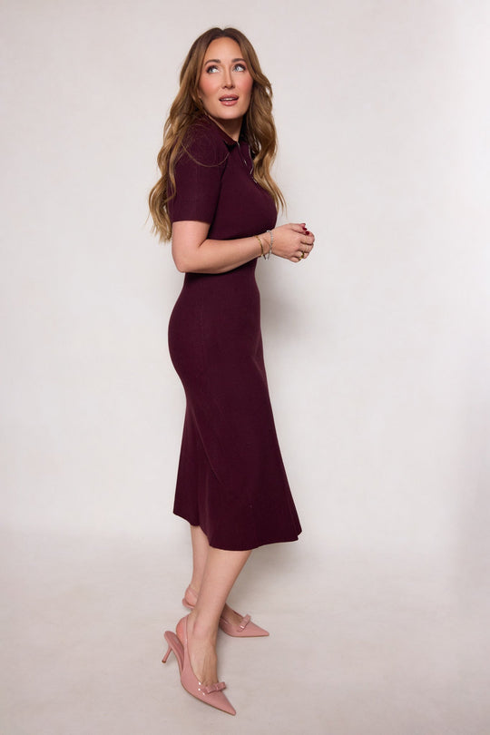 A woman with long wavy hair, wearing the Ivy City Co Ora Dress in Burgundy—a short-sleeved midi dress with a flowy pencil skirt—and pink heels, stands against a plain light background, smiling and gazing slightly to the side.