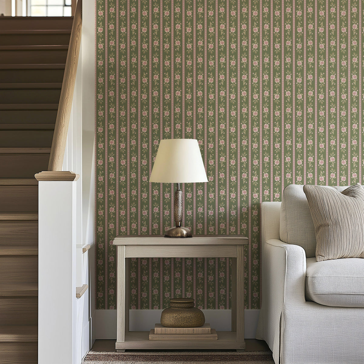 A beige table lamp sits on a wooden side table beside a light gray sofa, in front of striking Felicity Floral Wallpaper by Loomwell Home Goods, and next to a wooden staircase with white railings.