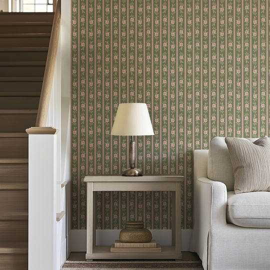 A beige table lamp sits on a wooden side table beside a light gray sofa, in front of striking Felicity Floral Wallpaper by Loomwell Home Goods, and next to a wooden staircase with white railings.