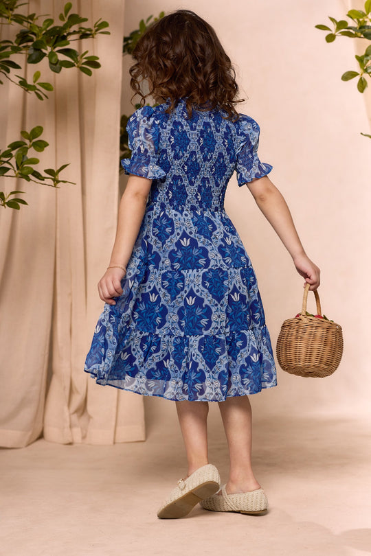 A young girl wearing the Ivy City Co Mini Cordelia Dress in Trellis Foulard stands barefoot on tiptoe, facing away while holding a small wicker basket. Green leaves and beige curtains frame the scene.